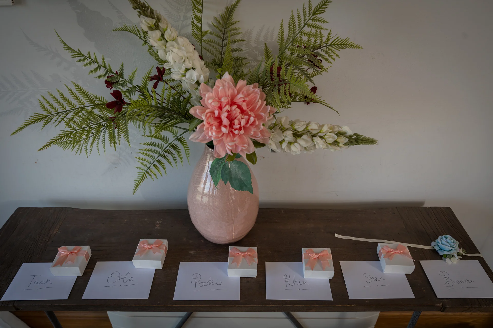 A rustic wooden table displays place cards with handwritten names alongside small white favor boxes tied with pink ribbons, beneath a ceramic vase filled with fern fronds, white snapdragons, and a central pink dahlia.