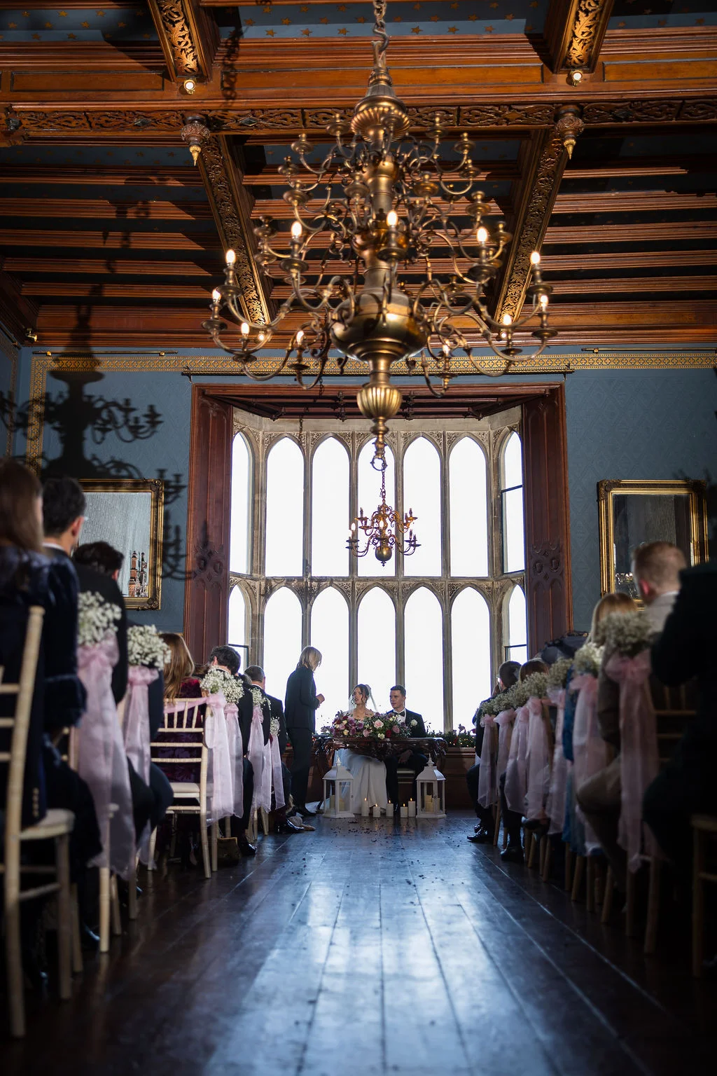 A bride and groom are seated at a table during an indoor wedding ceremony in a grand, wood-panelled historic room featuring large arched windows and an ornate chandelier.
