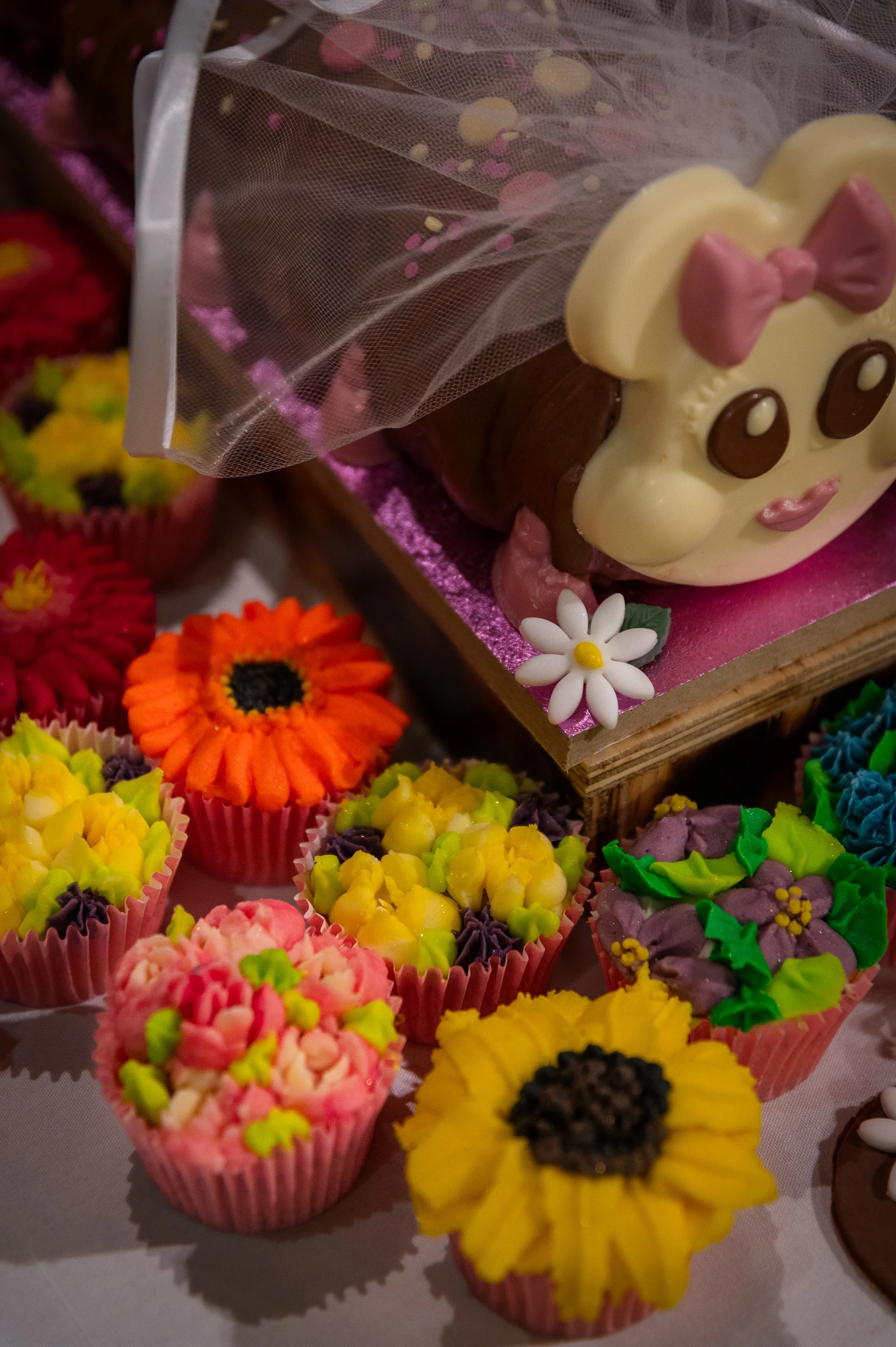 A whimsical novelty caterpillar cake draped in a miniature bridal veil is surrounded by an assortment of brightly coloured floral cupcakes.