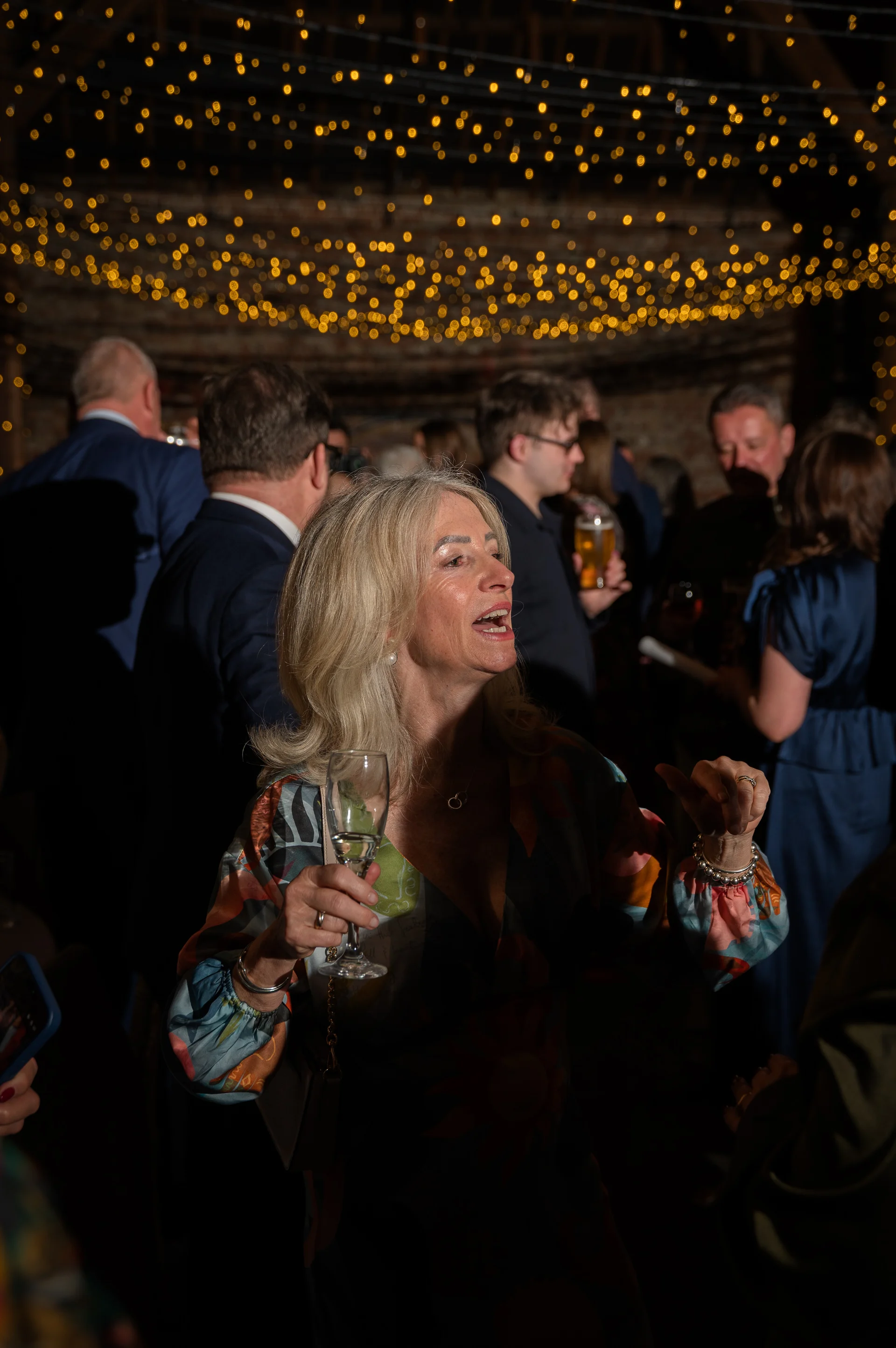 A wedding guest holds a glass of champagne while enjoying the evening party beneath a glowing canopy of fairy lights inside a rustic barn.