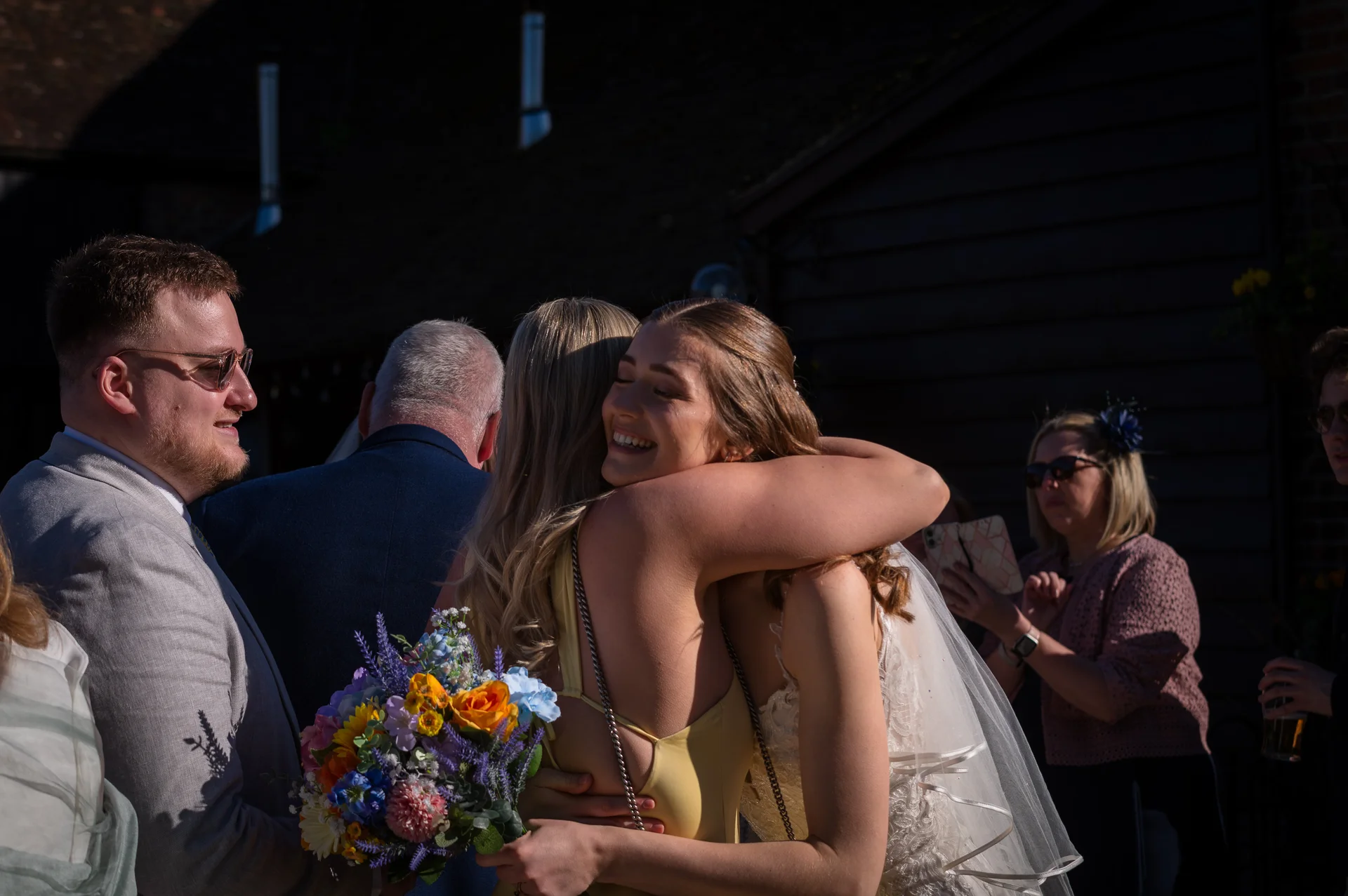 A guest in a yellow dress warmly embraces the smiling bride outdoors in the bright golden sunshine while holding a colourful bouquet.