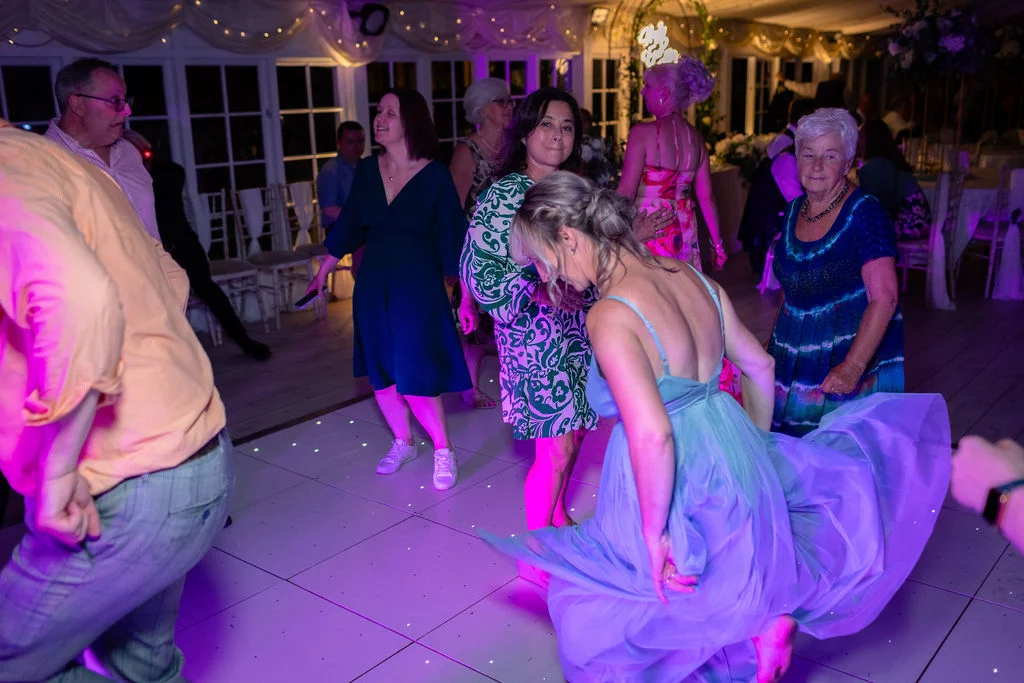 Wedding guests enjoy the evening reception on a glowing dance floor under purple lighting while a woman twirls her blue dress.