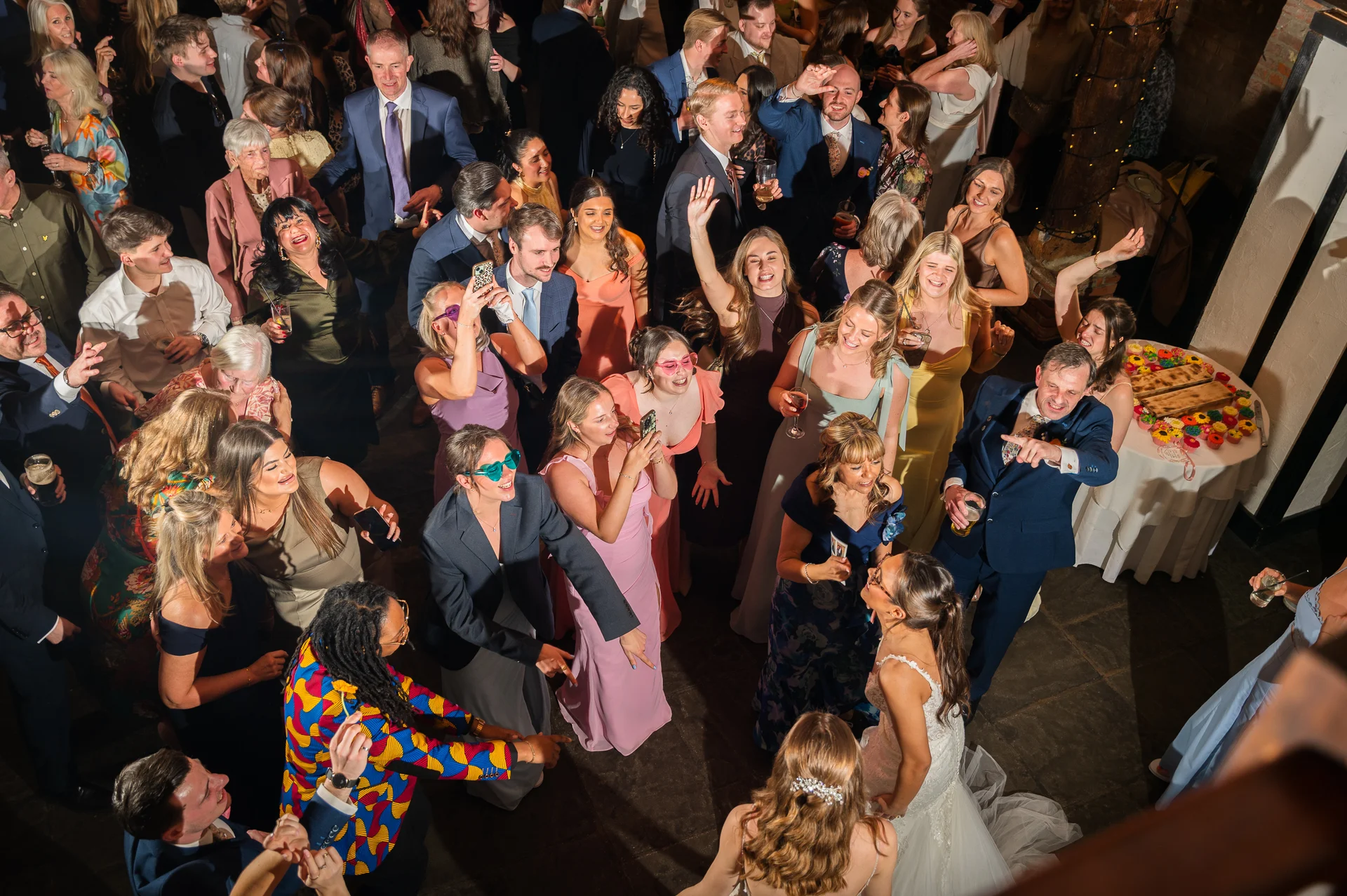 An overhead shot captures a lively, crowded dance floor filled with wedding guests laughing, drinking, and dancing around the bride and groom.