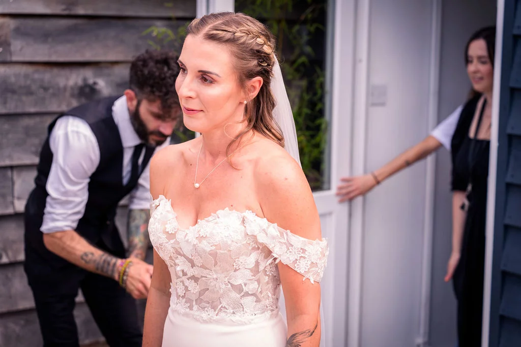 A glowing bride stands in a lace off-the-shoulder wedding gown while a groom adjusts her attire in the background.