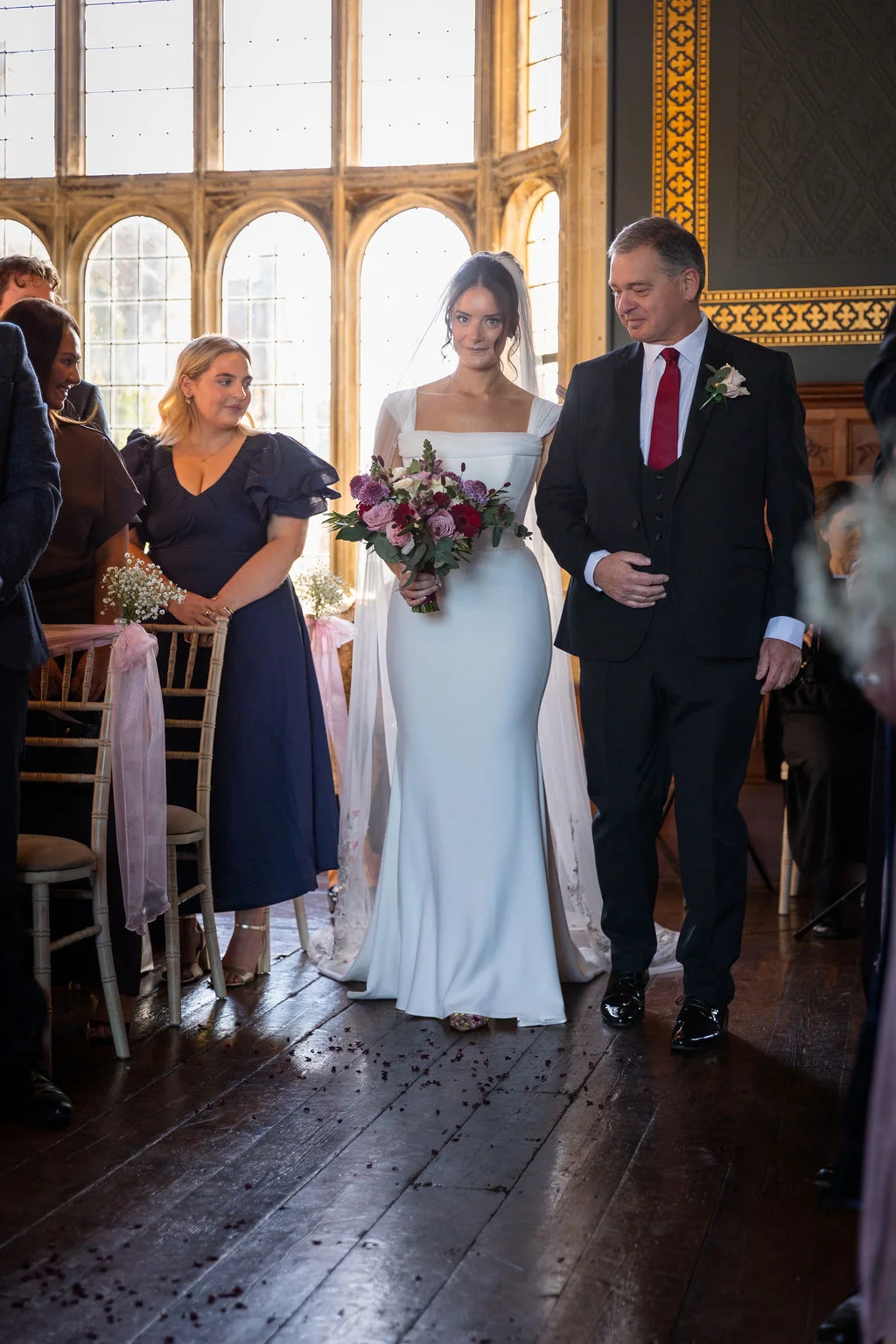 A bride walks down the aisle arm in arm with her father inside a grand hall with large windows, holding a colourful bouquet.