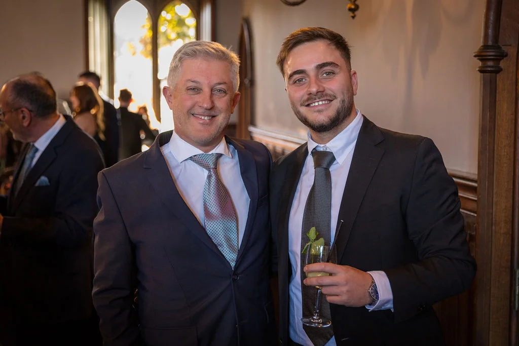 Two smiling male wedding guests in smart suits pose together indoors during the drinks reception, with one holding a garnished cocktail glass.