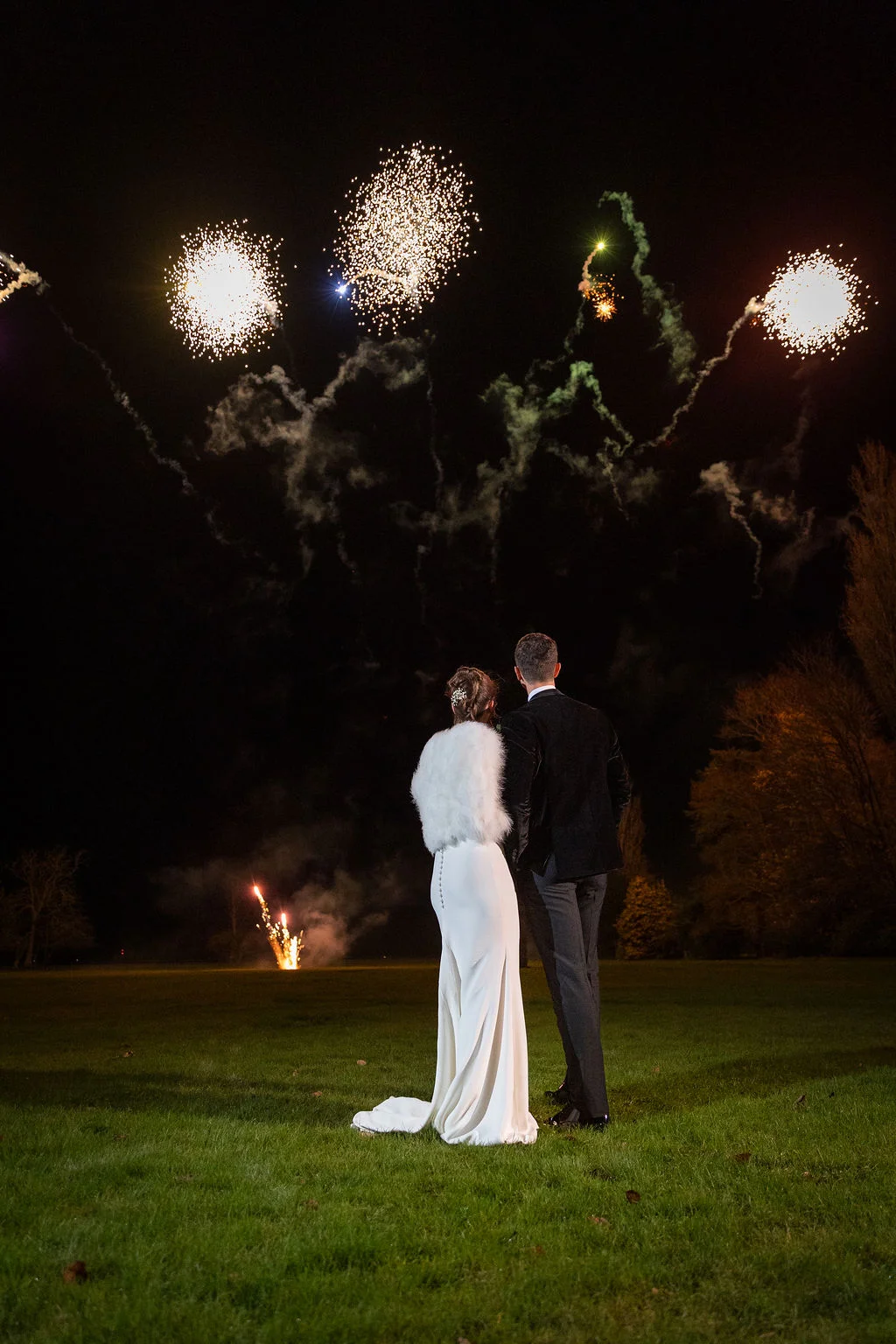 A newlywed couple stands on a lawn at night, watching a vibrant fireworks display illuminate the dark sky above them.