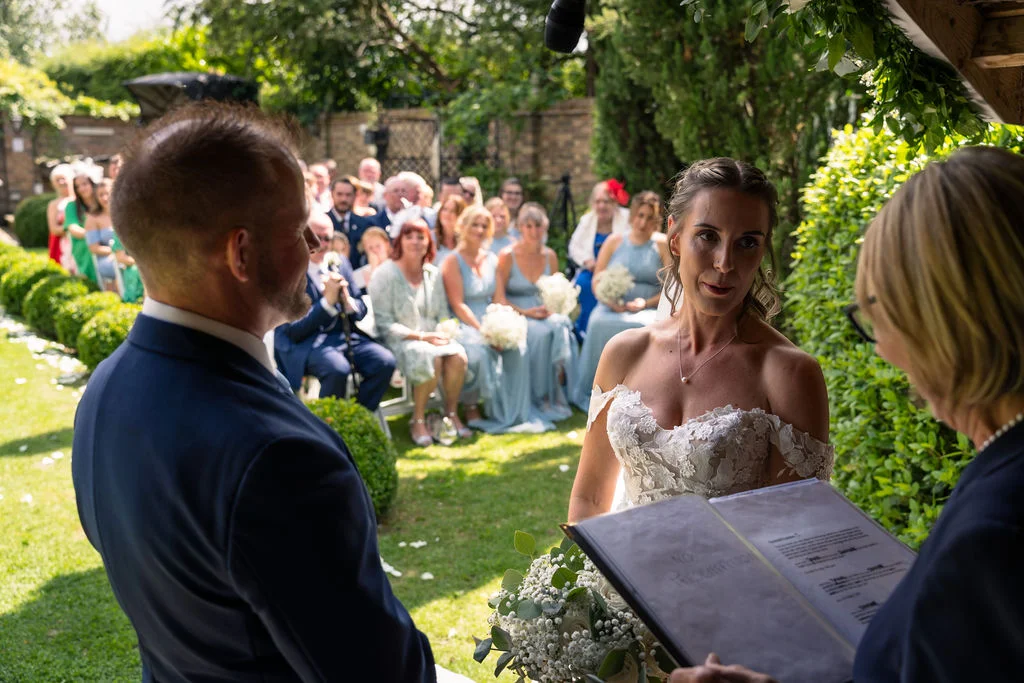 An outdoor wedding ceremony takes place in a lush garden, with the bride and groom standing before the celebrant as seated guests watch on.