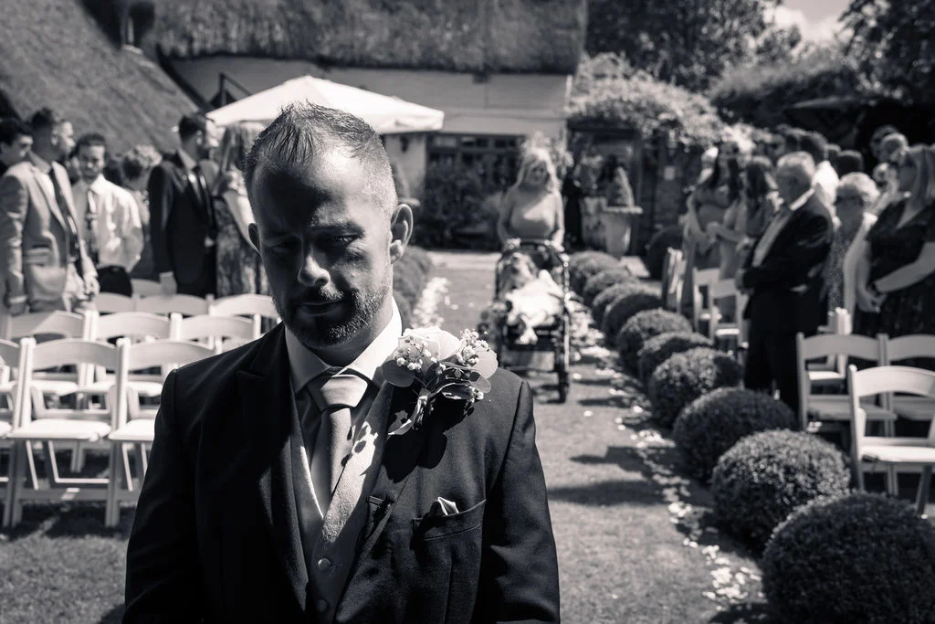 A groom in a three piece suit with a buttonhole stands outdoors waiting as guests are seated along the aisle.