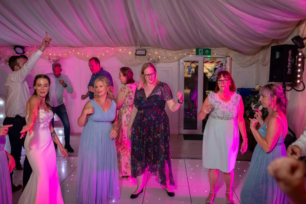 A lively group of wedding guests and the bride dance energetically on an illuminated dance floor beneath a draped marquee ceiling with pink lighting.