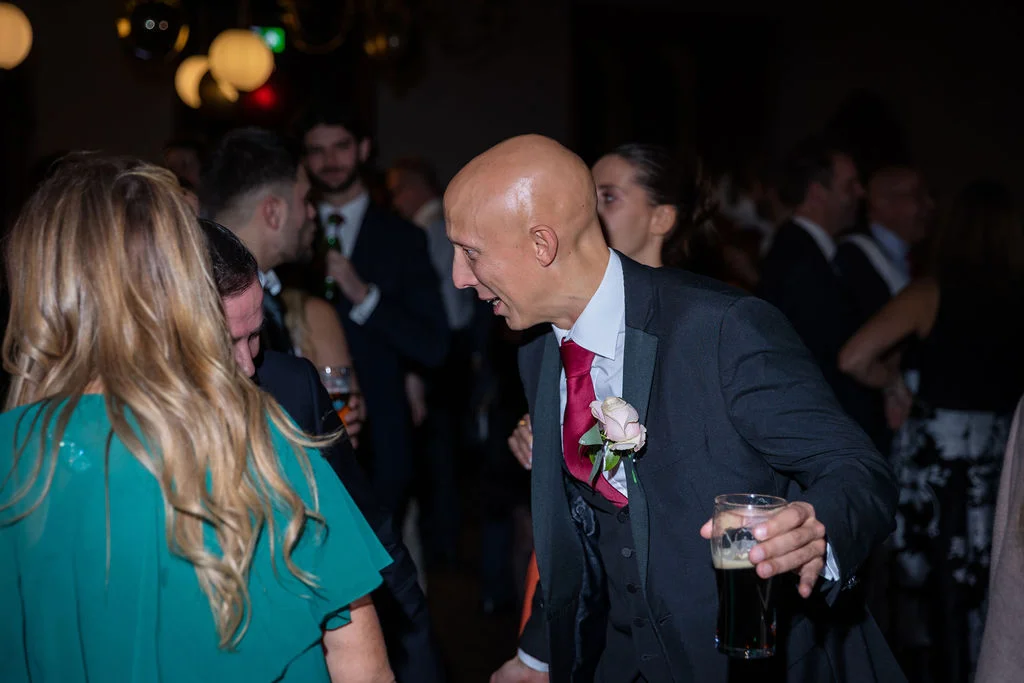 A guest in a black tuxedo and red tie laughs while holding a pint of beer during a lively evening wedding reception.