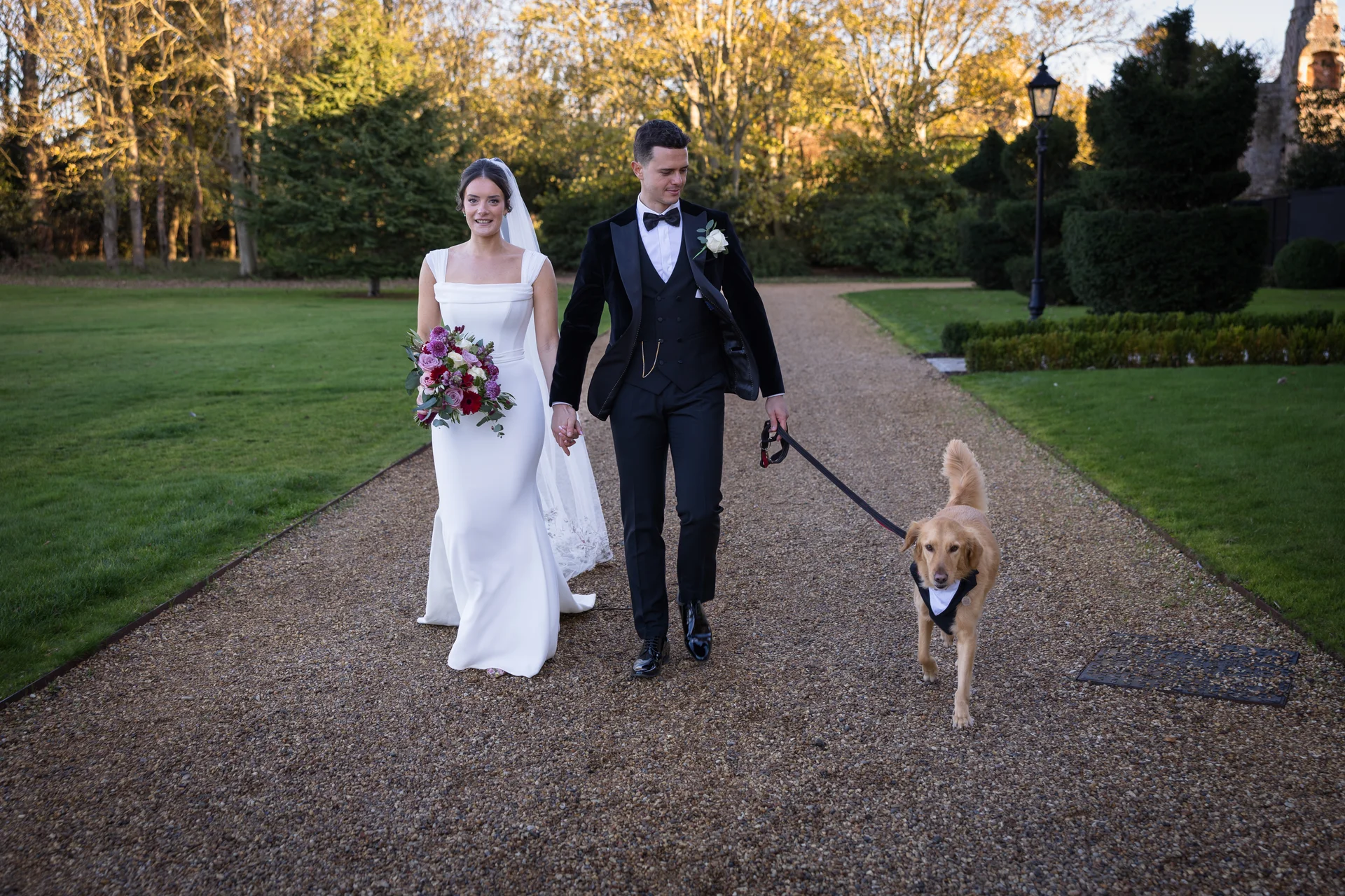 A newlywed couple smiling as they walk hand in hand along a gravel path in golden hour sunlight, accompanied by their golden retriever wearing a bow tie.