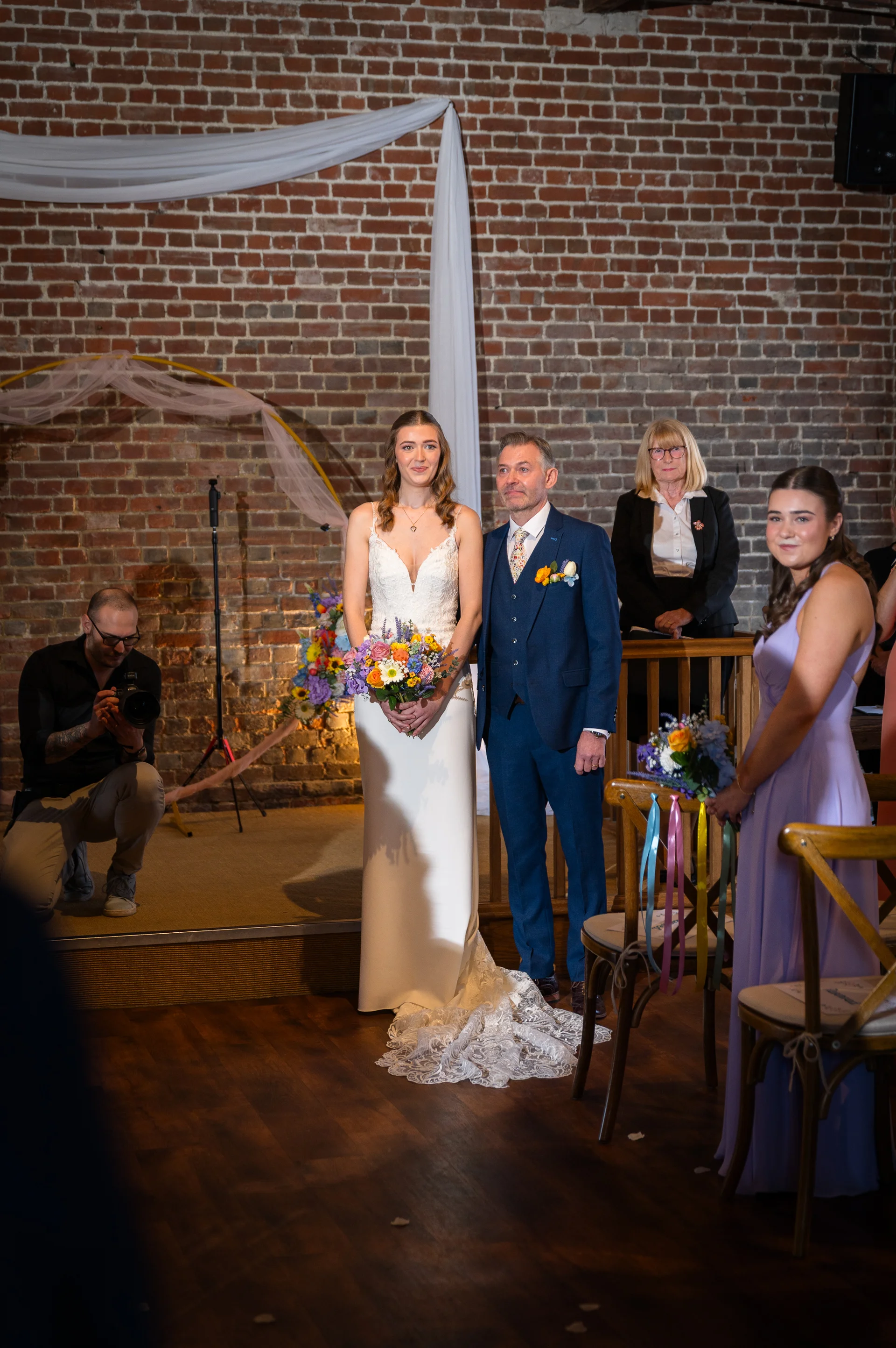 A bride in a white column gown holding a colourful bouquet stands with her groom in a blue suit during a barn wedding ceremony.
