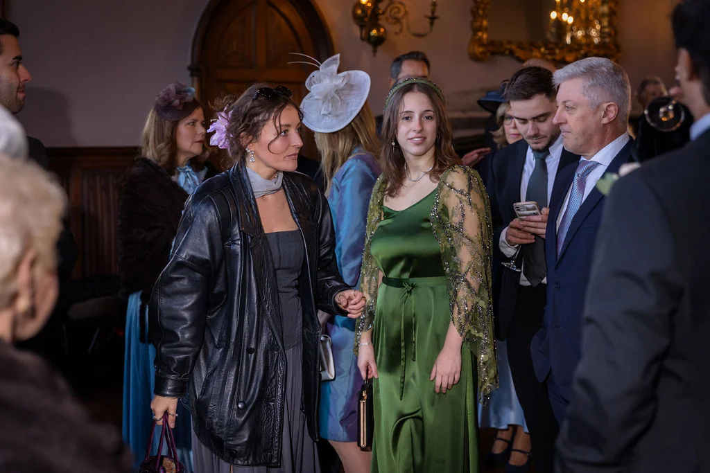 A candid photograph capturing wedding guests mingling inside a manor house, featuring a guest in a flowing emerald green satin dress with beaded capelet and another in a black leather jacket.