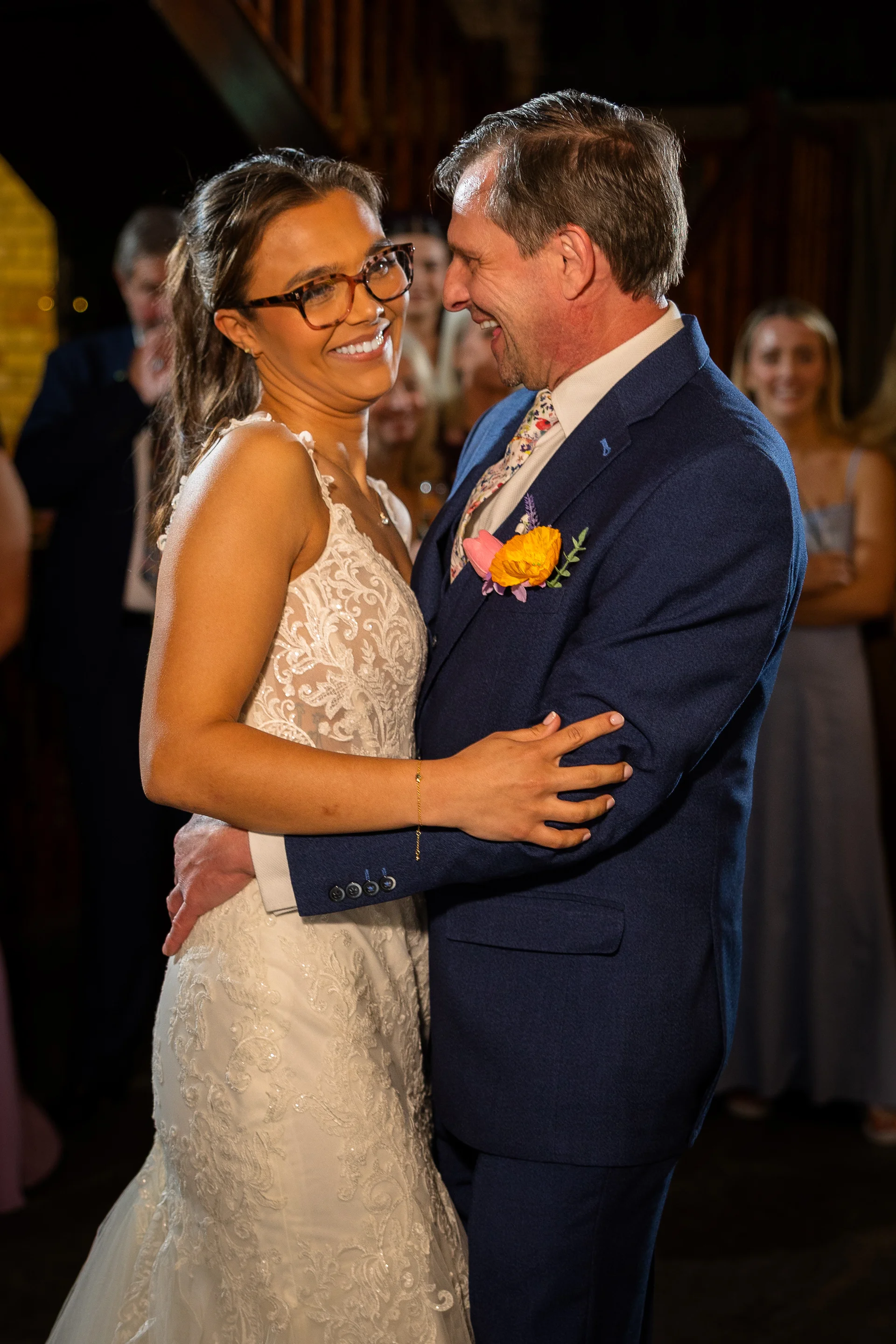 A smiling bride wearing glasses and a lace wedding dress embraces her groom during their first dance, with wedding guests watching happily in the background.