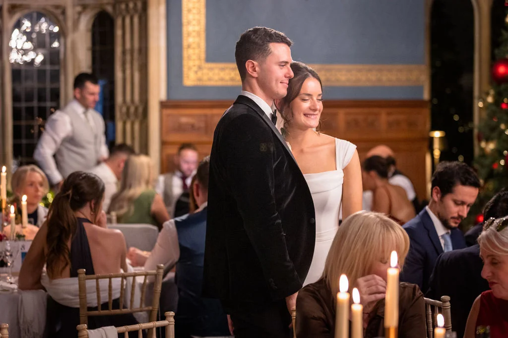 Newlyweds stand together smiling during their wedding breakfast inside a grand candlelit manor house dining room surrounded by seated guests.