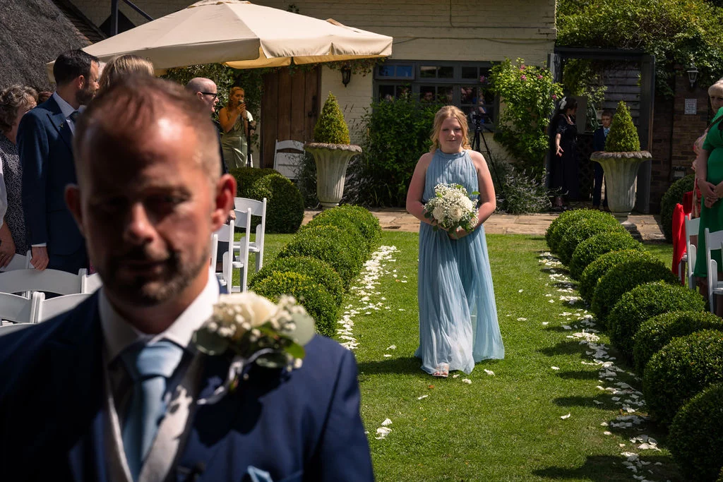 A groom stands in the foreground watching as a bridesmaid in a dusty blue gown walks down the garden aisle lined with topiary and white petals.