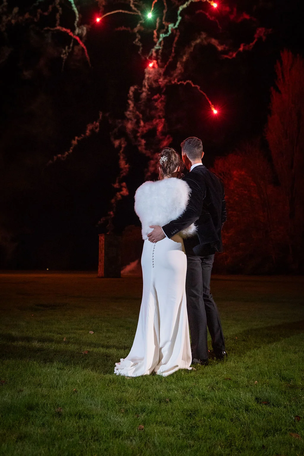 A newlywed couple stand on a lawn at night, embracing while watching colourful fireworks light up the sky above them.