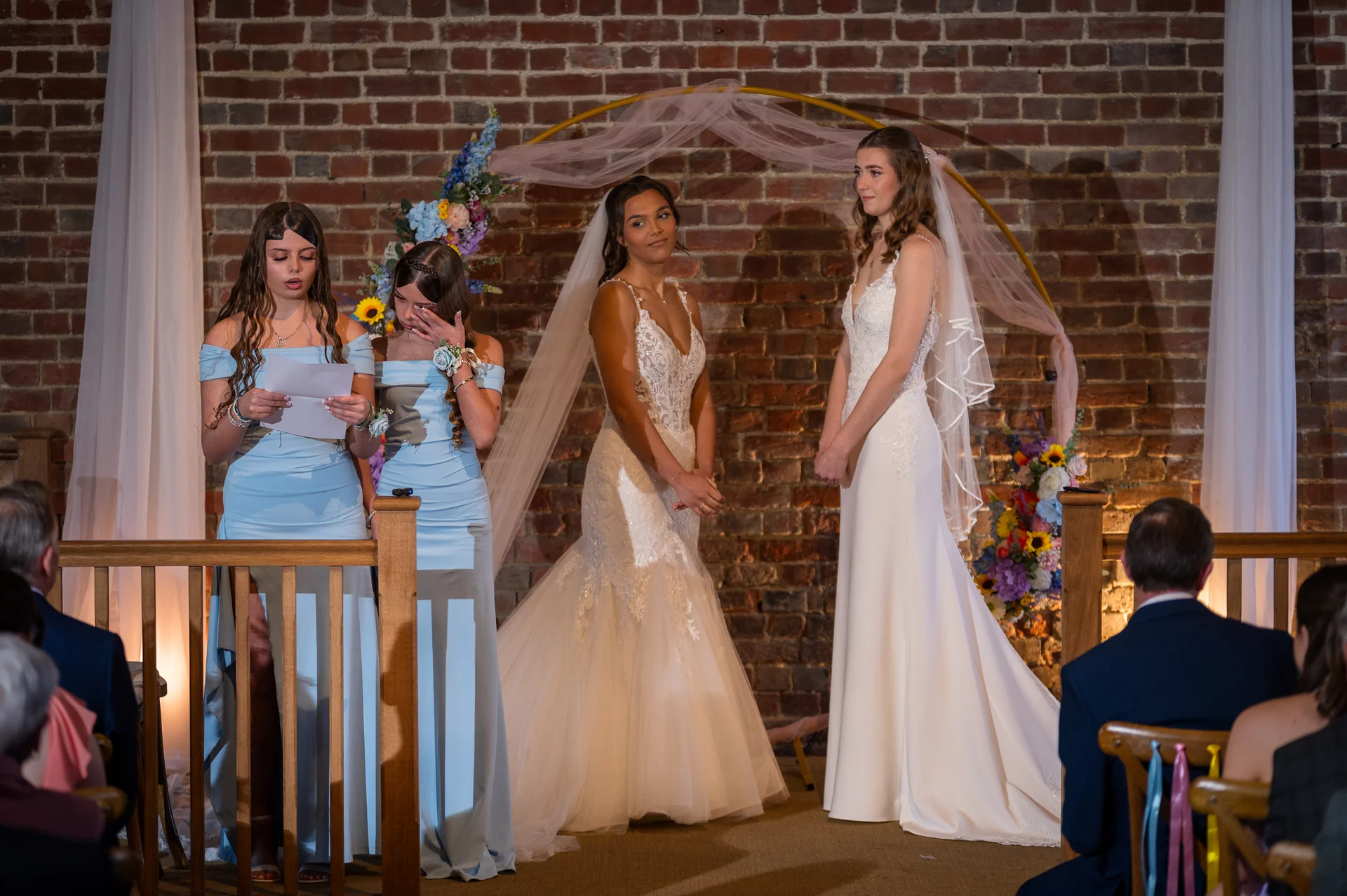 Two brides stand together at the altar during their emotional wedding ceremony, while bridesmaids in light blue dresses look on near an exposed brick backdrop.