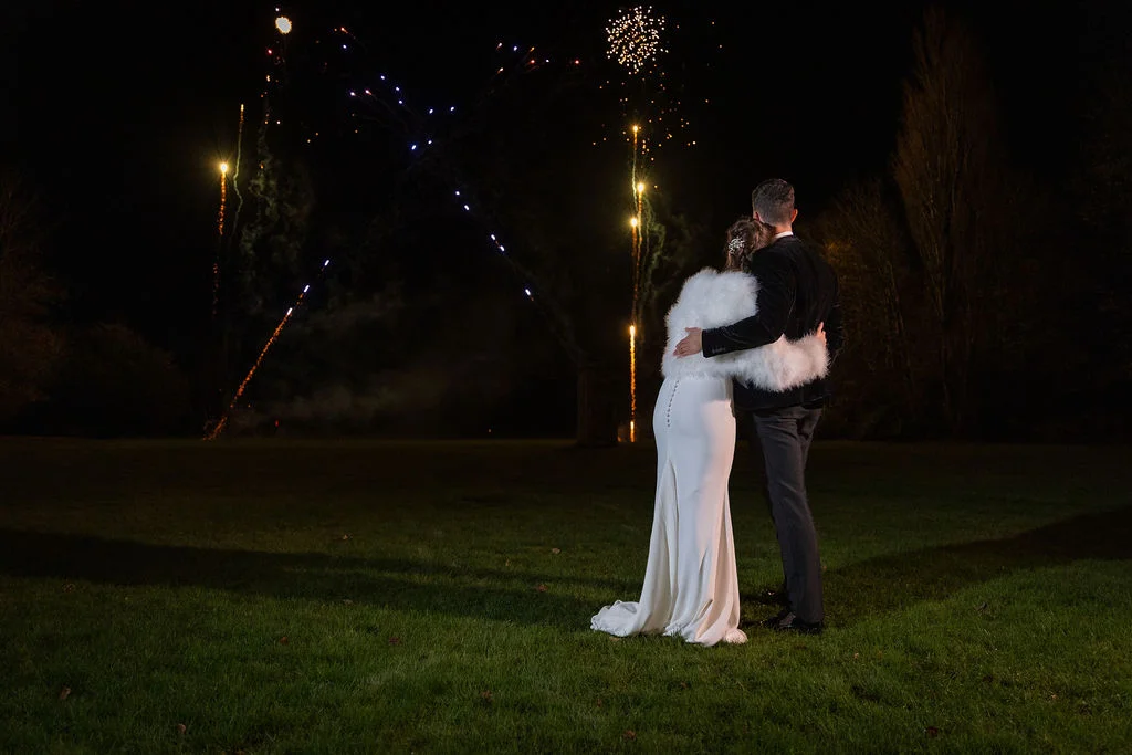 A newlywed couple embraces outdoors on a grassy lawn at night, watching a vibrant fireworks display light up the dark sky.