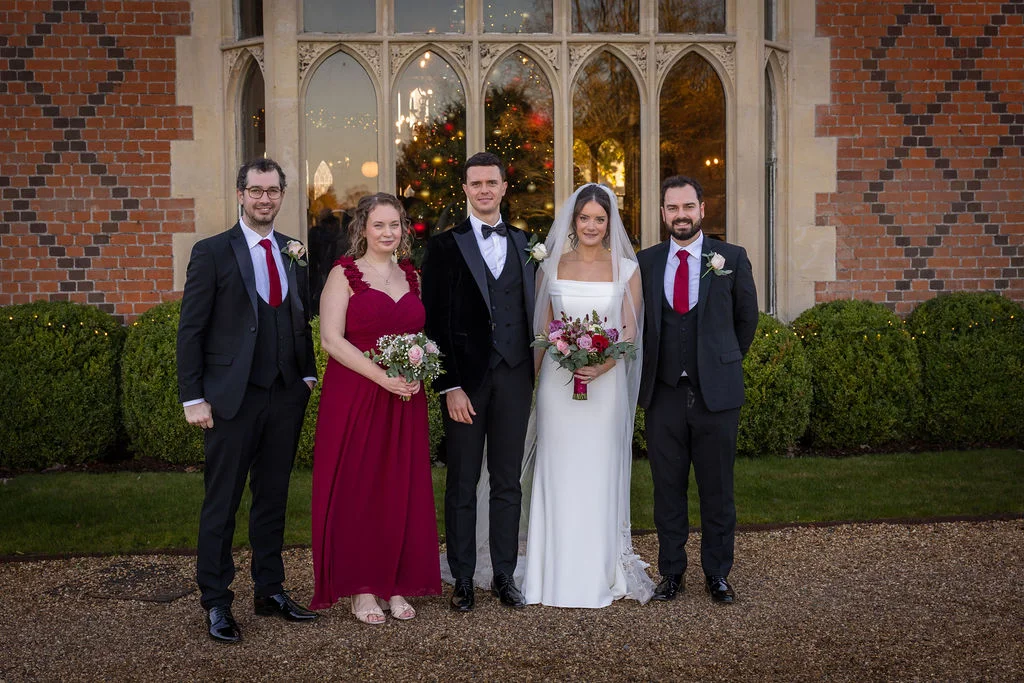 A bridal party featuring a bride in a white gown and veil, a groom in a black velvet tuxedo, alongside groomsmen in black suits with red ties and a bridesmaid in a red dress, pose outdoors in front of a historic brick building with large arched windows.
