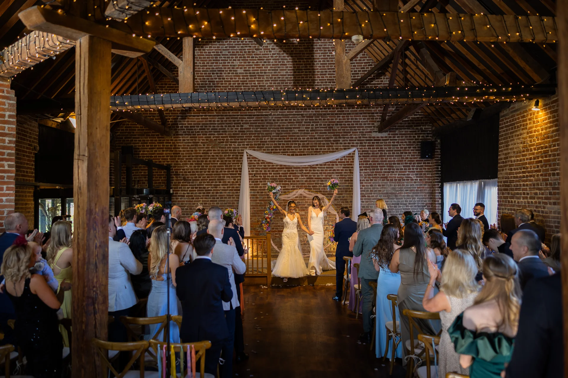 Two brides celebrate raising their bouquets as they walk back down the aisle together inside a rustic brick barn adorned with fairy lights.