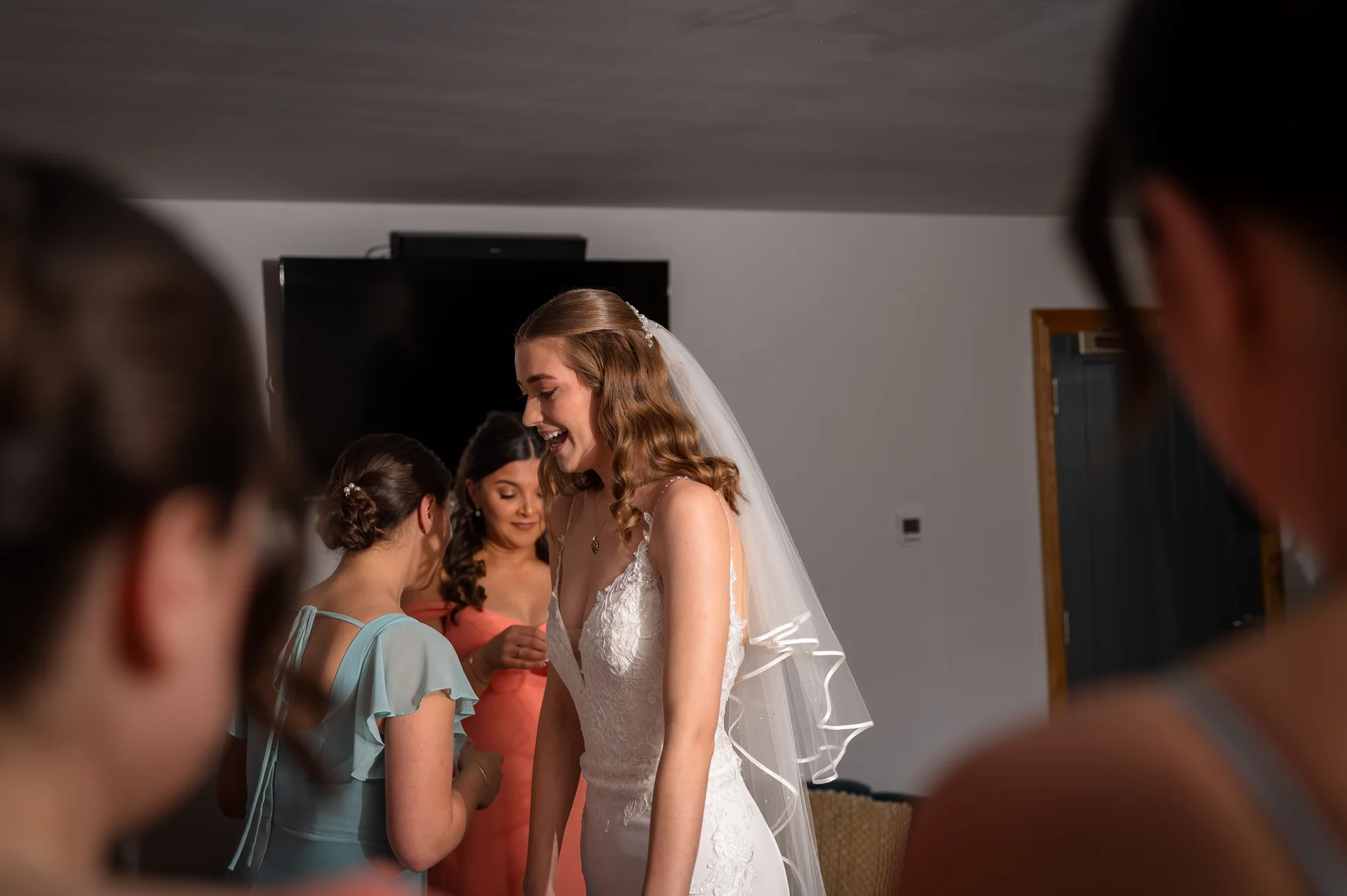 A laughing bride in a lace v-neck wedding dress and fingertip veil shares a joyful moment during bridal prep while bridesmaids look on in anticipation.