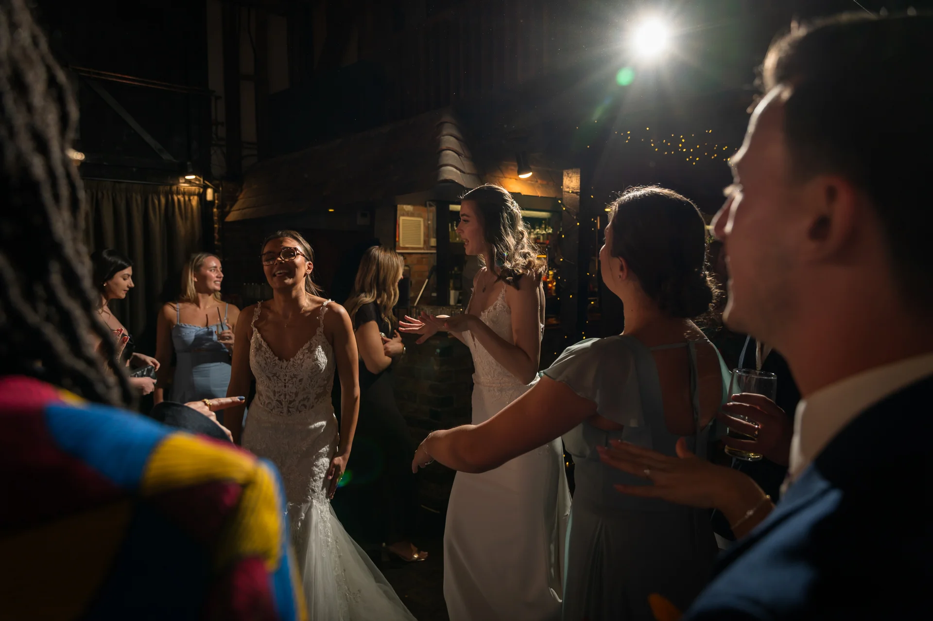 Brides and wedding guests enjoying themselves on the dance floor during a lively evening reception illuminated by dramatic backlighting and string lights.