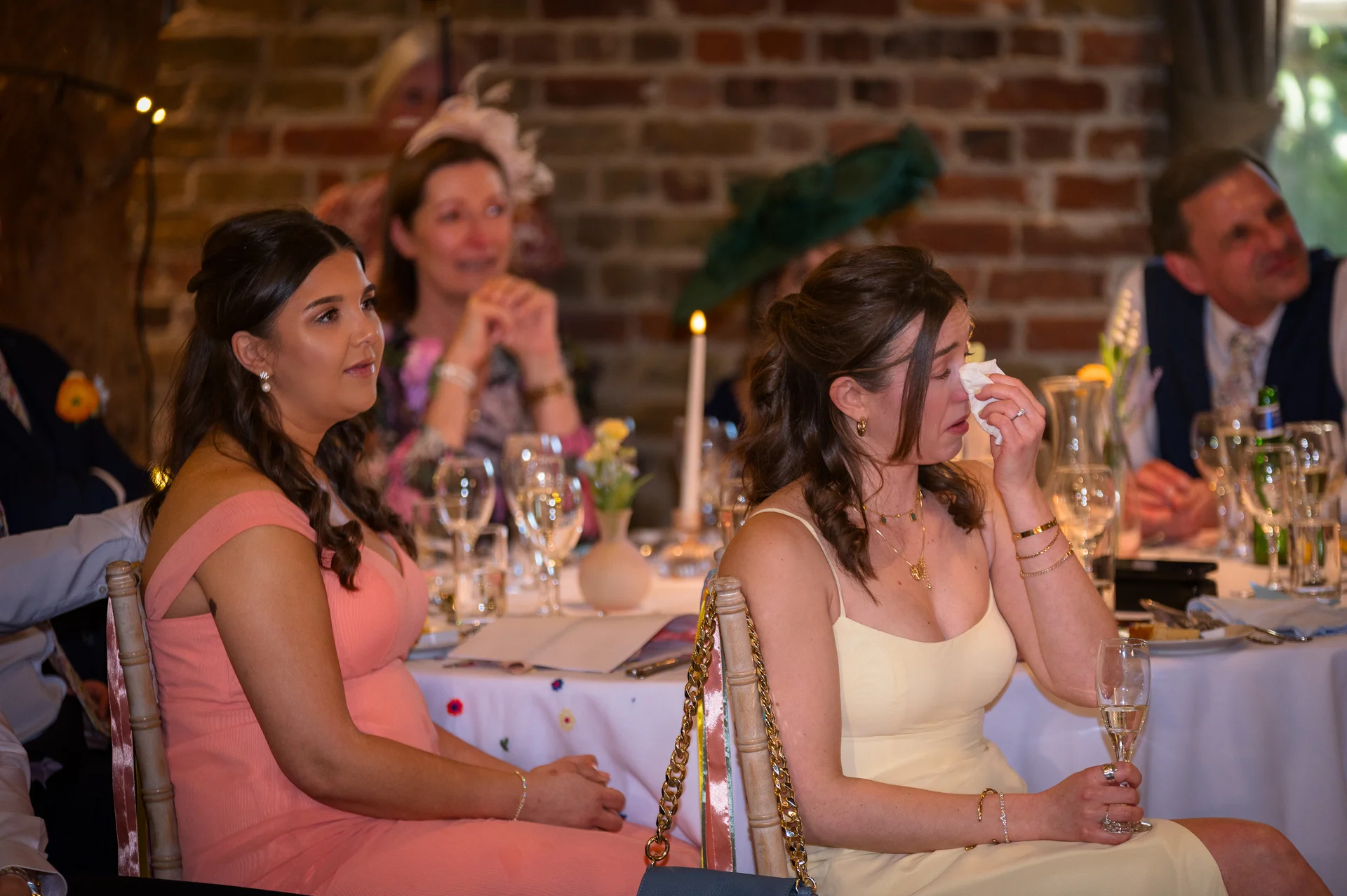 An emotional guest at a wedding reception wipes away a tear with a tissue during the speeches while seated at a candlelit banquet table.