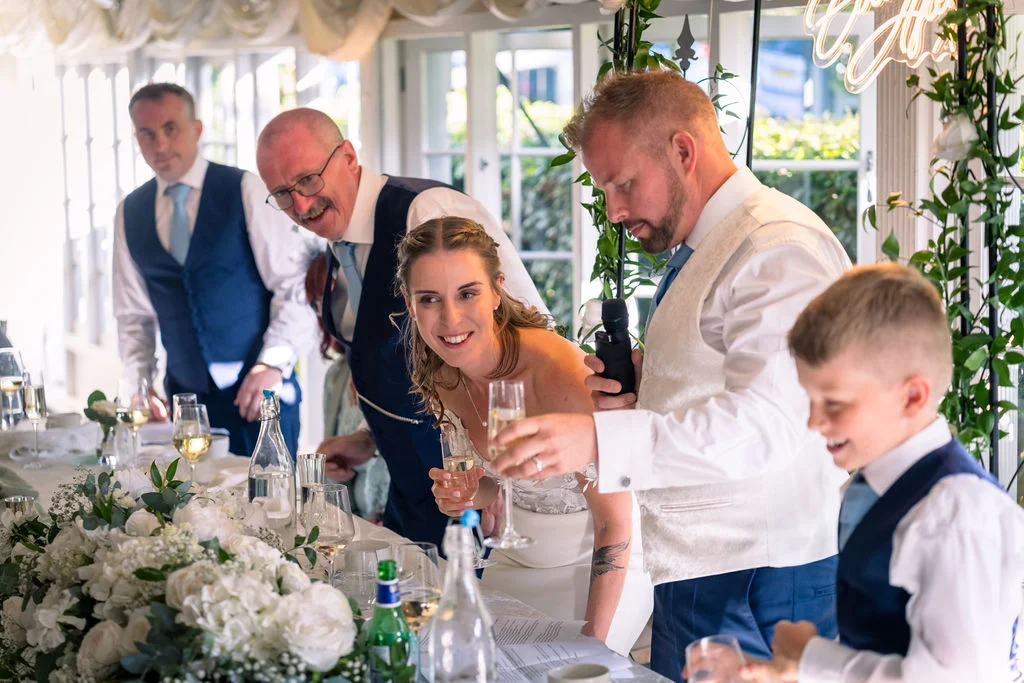 A smiling bride and a groom delivering a speech at a wedding reception table adorned with lush white floral runners and glassware.