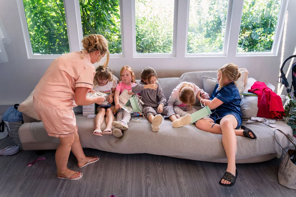 An adult in peach pyjamas stands by a sofa while several young girls and bridesmaids in matching sleepwear unwrap gifts in a bright, sunlit room.