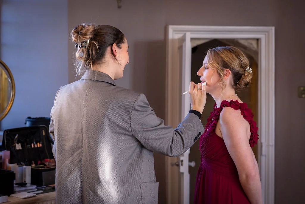A makeup artist applies finishing touches to a bridesmaid wearing a striking burgundy dress inside a brightly lit room.