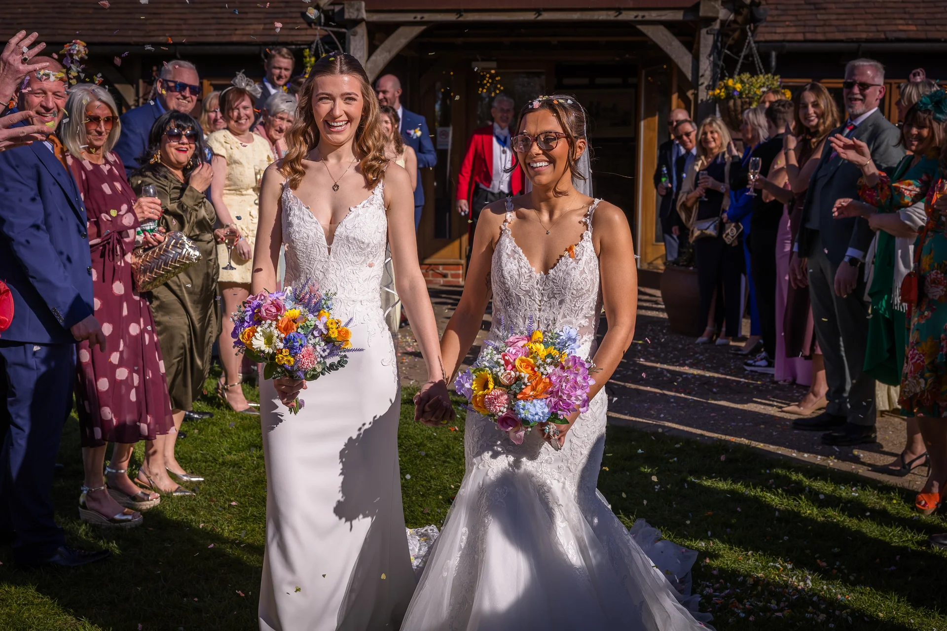 Two smiling brides holding hands and carrying colourful bouquets walk through a shower of confetti thrown by cheering wedding guests outdoors on a sunny day.
