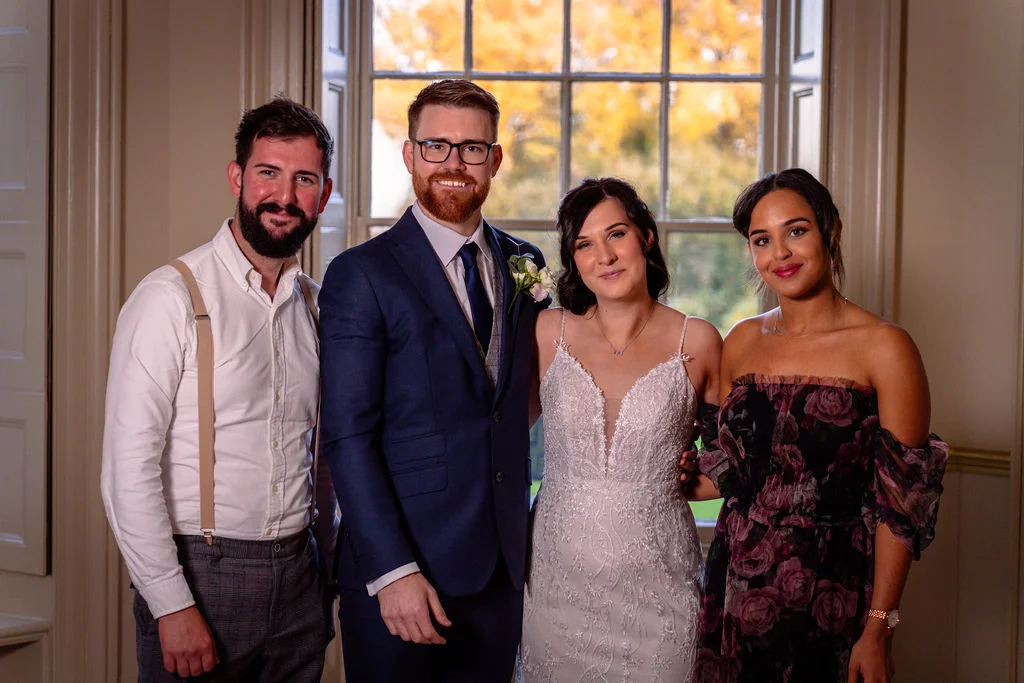 A newlywed couple and two members of their wedding party pose together indoors in front of a large window displaying autumn foliage.