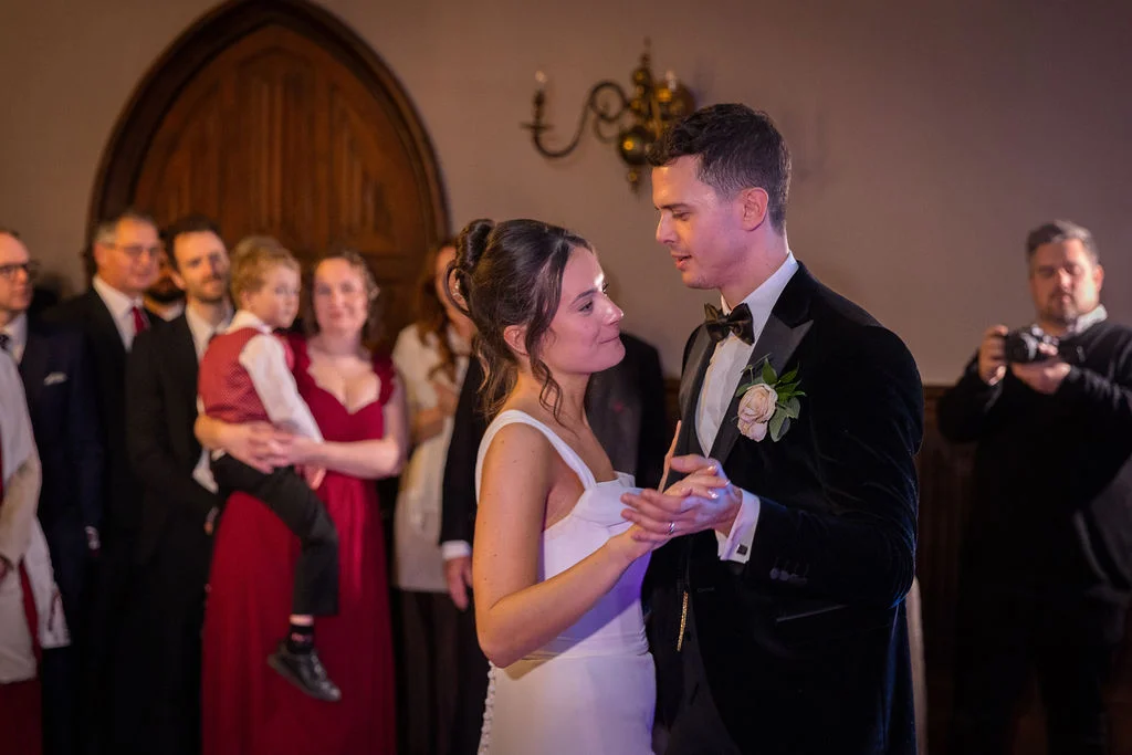 The newly married couple share a tender first dance together in a warm room surrounded by watching wedding guests.