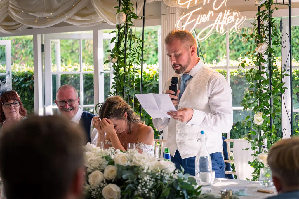 A groom delivers a speech while standing at the top table in a light-filled orangery decorated with white floral arches, causing his bride to cover her face and laugh.