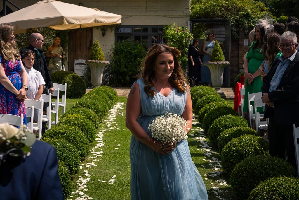 A bridesmaid in a flowing dusty blue dress walks down a garden aisle flanked by neat topiary bushes and scattered petals, carrying a gypsophila bouquet.