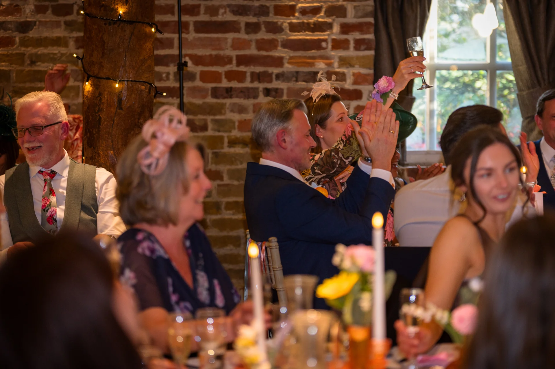 Wedding guests laugh and cheer enthusiastically during the reception speeches, raising a glass of champagne in an exposed brick venue.