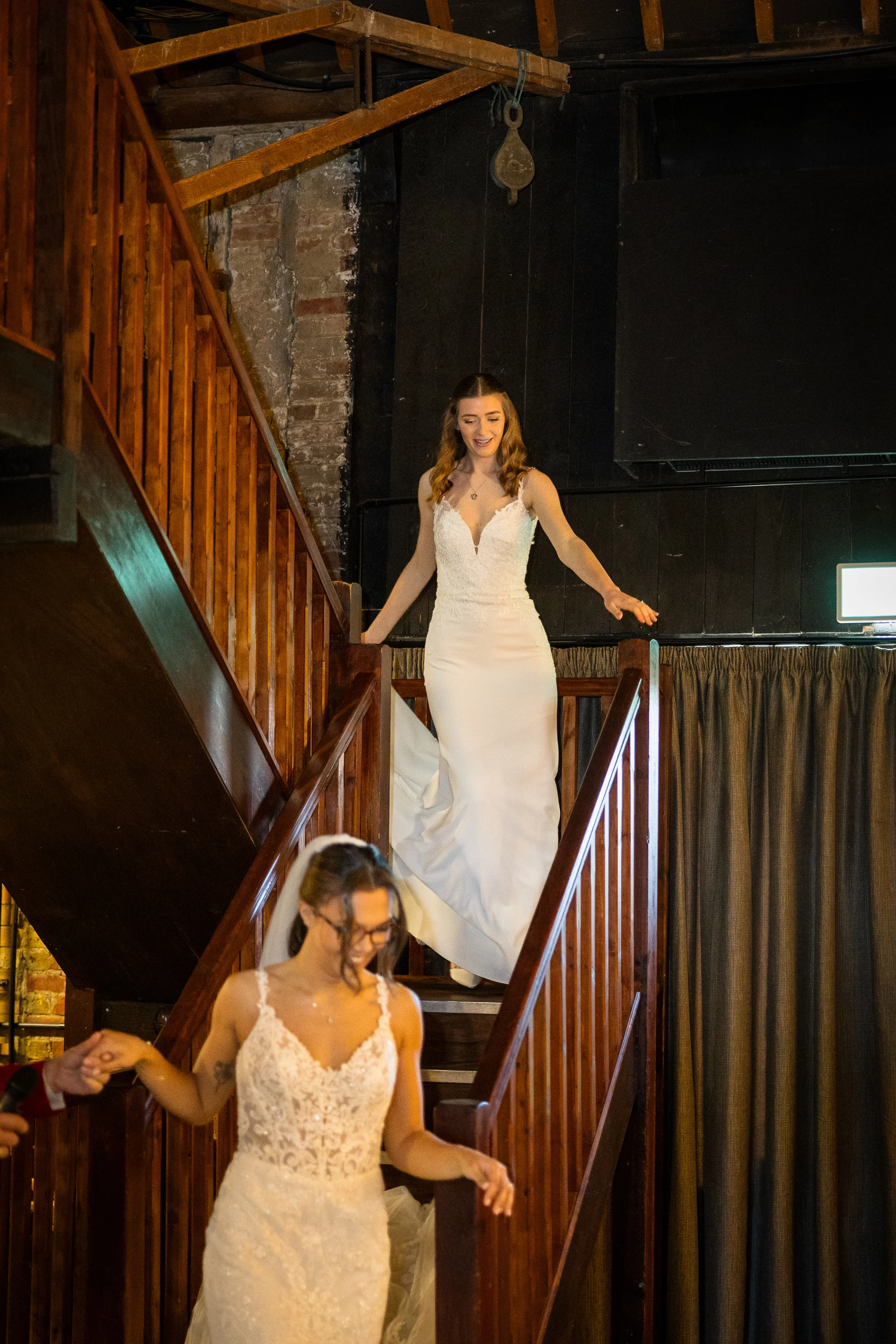 Two smiling brides in elegant white wedding dresses walk down a rustic wooden staircase inside a converted barn venue.