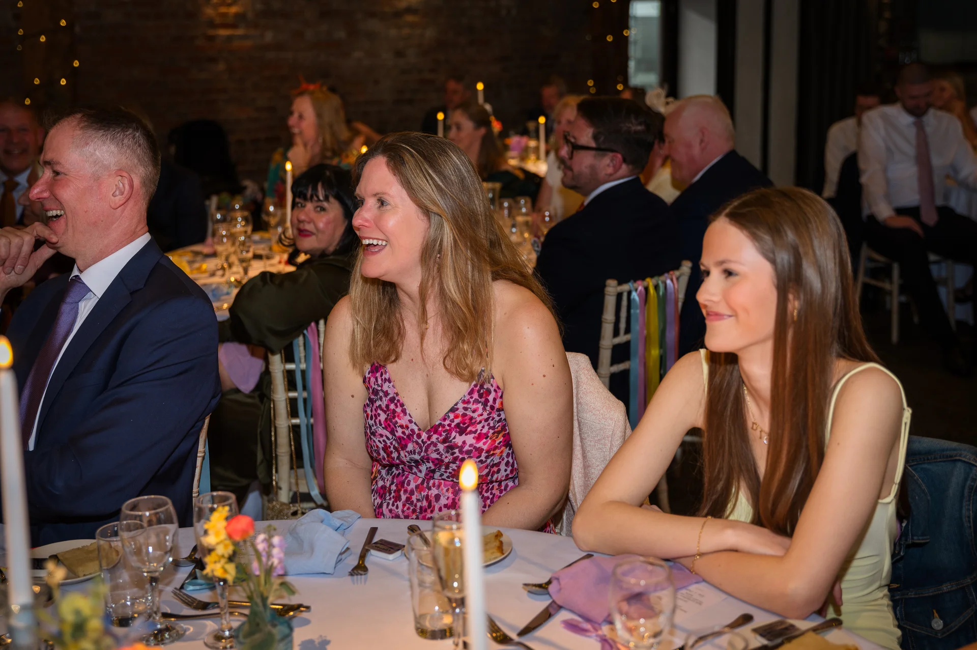 Wedding guests laugh joyfully during speeches at a candlelit indoor reception table featuring exposed brickwork.
