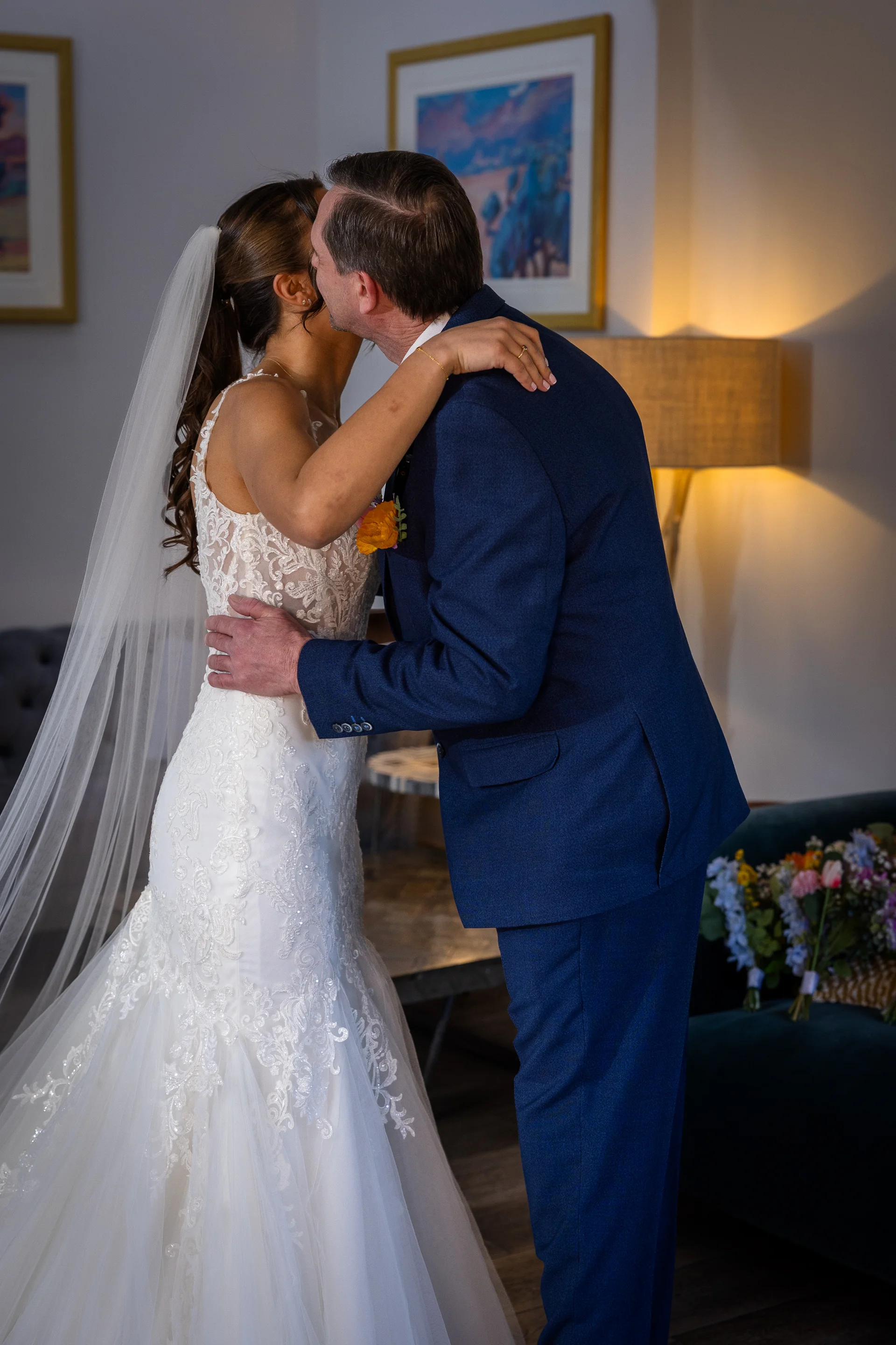 A bride in a lace a-line gown and veil shares a tender embrace and kiss with her groom in a navy suit inside a cozy hotel room.