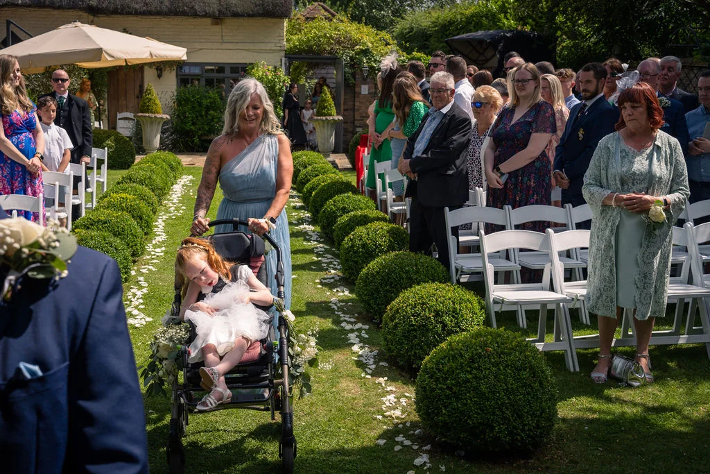 A young flower girl in a wheelchair is pushed down a sunny garden aisle lined with neatly trimmed box hedges and scattered white petals, watched by seated wedding guests.