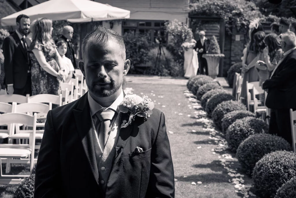 An emotional groom stands at the top of an outdoor aisle lined with clipped box hedge spheres, watching the bride approach during a garden ceremony.