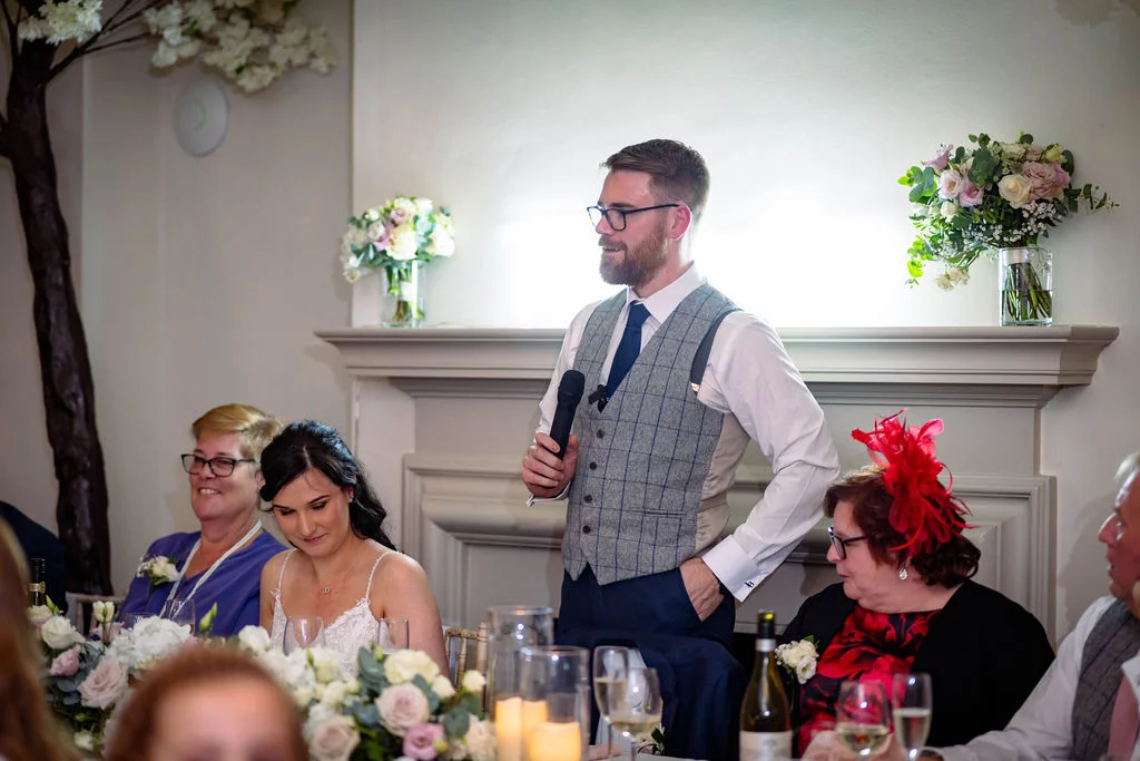 A groom stands to give a speech during the wedding reception, holding a microphone while wearing a tweed waistcoat, with the bride and guests seated nearby.