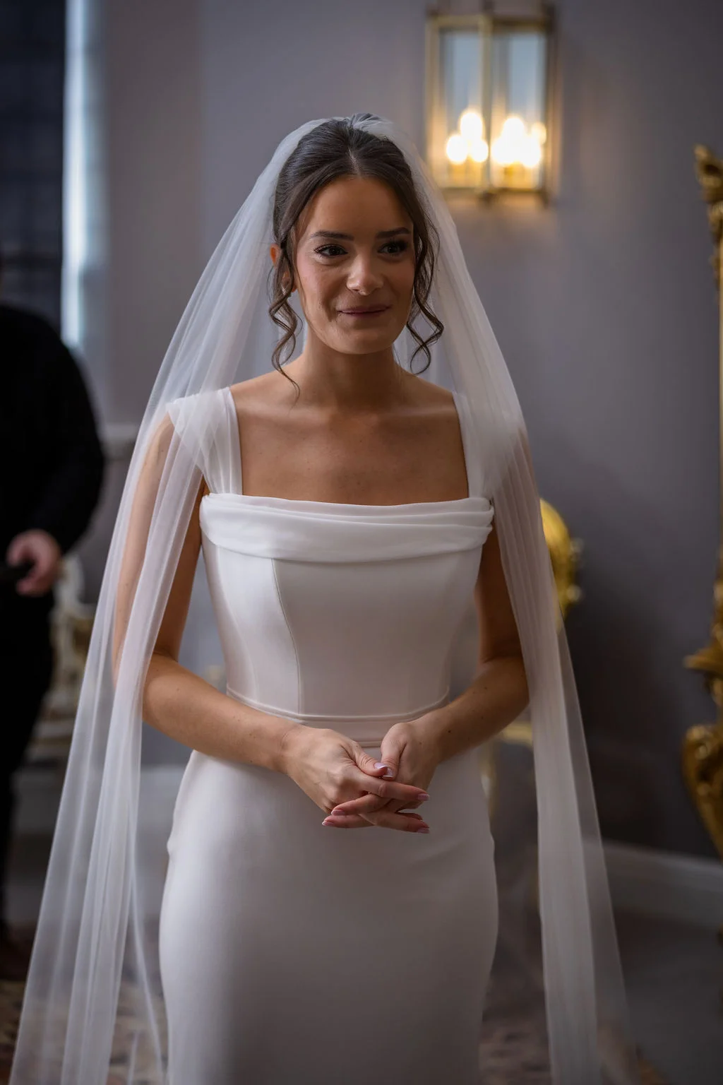 A stunning portrait of a bride smiling softly while wearing a sleek crepe column gown with a square neckline and a flowing chapel veil.