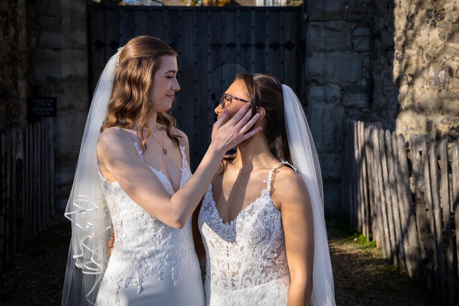 Two smiling brides in matching lace wedding dresses stand lovingly together in front of historic wooden doors during golden hour.