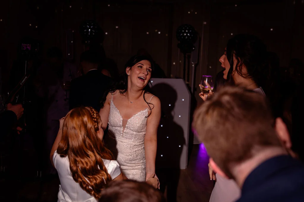 A joyful bride laughs while dancing with guests on a lively evening reception dance floor under atmospheric lighting.