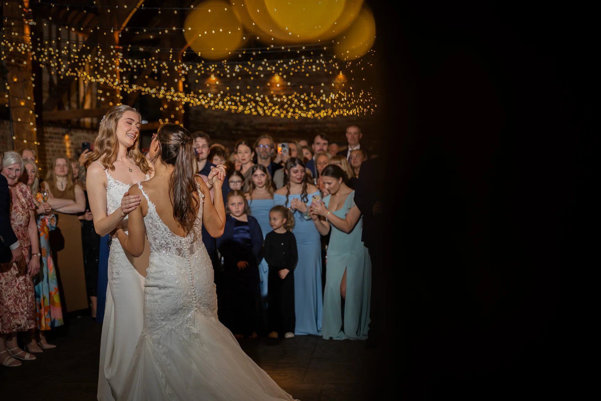 Two brides share a joyful first dance together under a glowing canopy of fairy lights as wedding guests watch from the background.