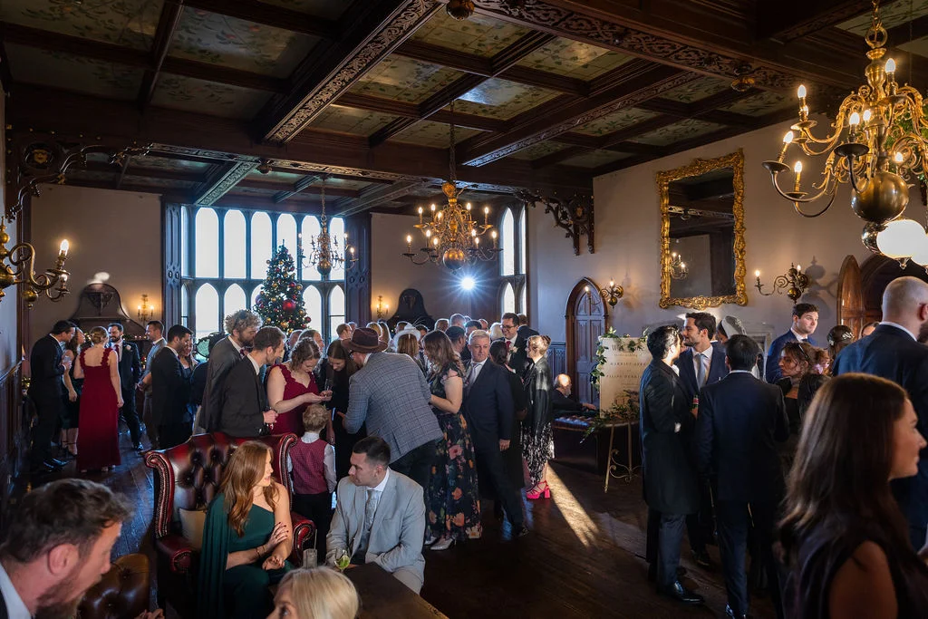 Wedding guests mingle during a lively drinks reception inside a historic wood-panelled hall adorned with ornate chandeliers and stained glass windows.