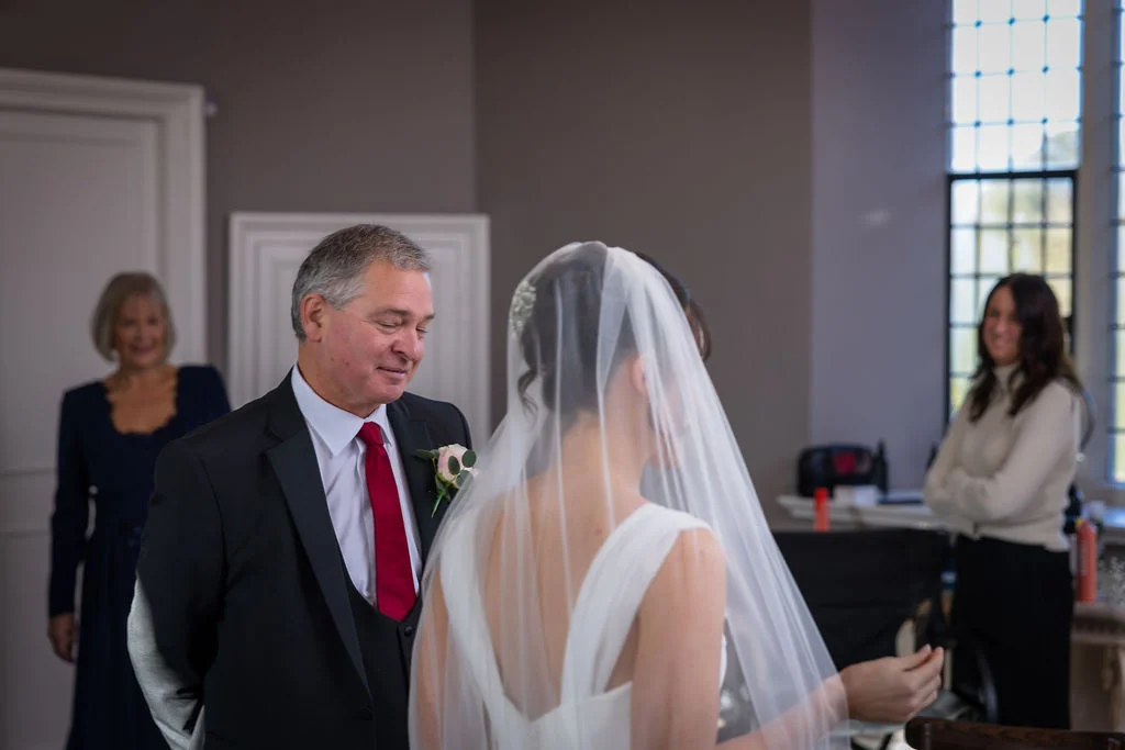 A father smiles warmly as he sees his daughter in her wedding dress and veil during a special getting ready moment.
